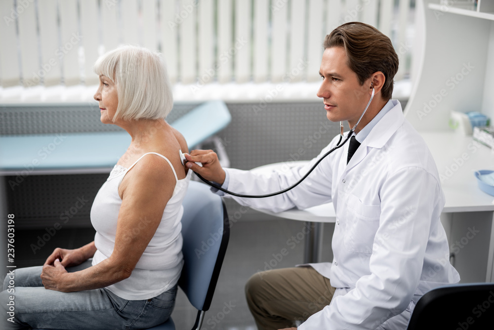 Fototapeta premium Calm aged woman sitting on the chair and professional general practitioner using stethoscope during the examination