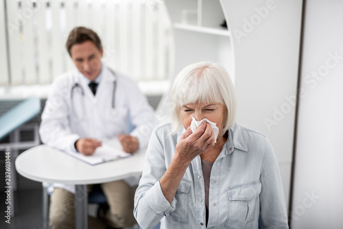 Elderly lady putting napkin to her nose and sneezing while the doctor sitting on the background