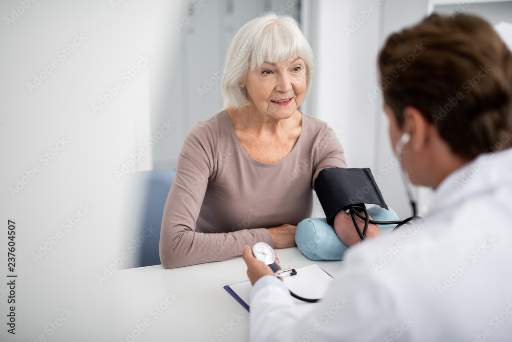 Fototapeta premium Relaxed elderly lady sitting in front of her practitioner and watching him measuring her blood pressure