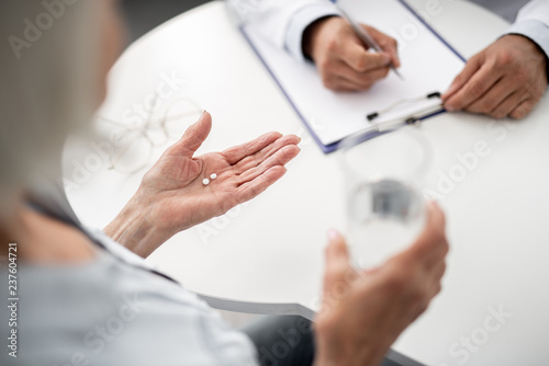 Elderly woman holding glass of water in right hand and two white pills in left hand while her doctor writing