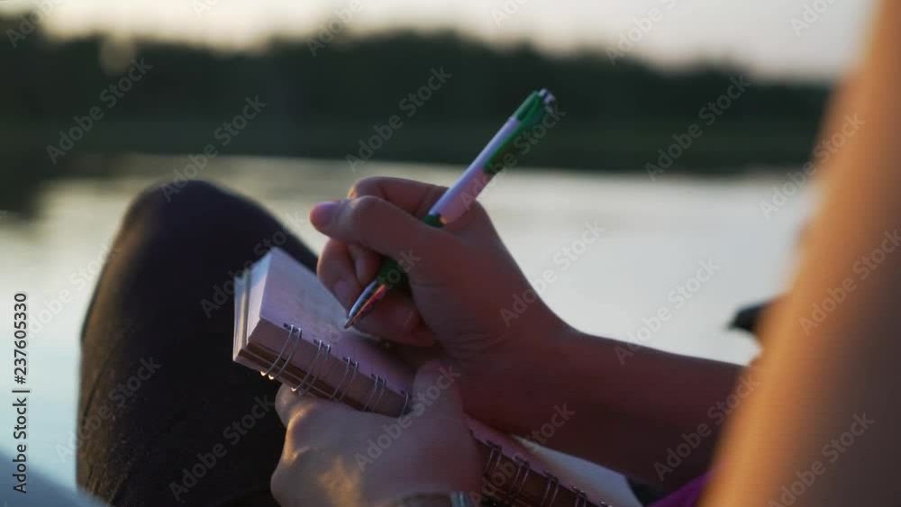 Close up of young white millenial woman journaling with pen while sitting in chair on pier overlooking beautiful scenic lake vista at sunset