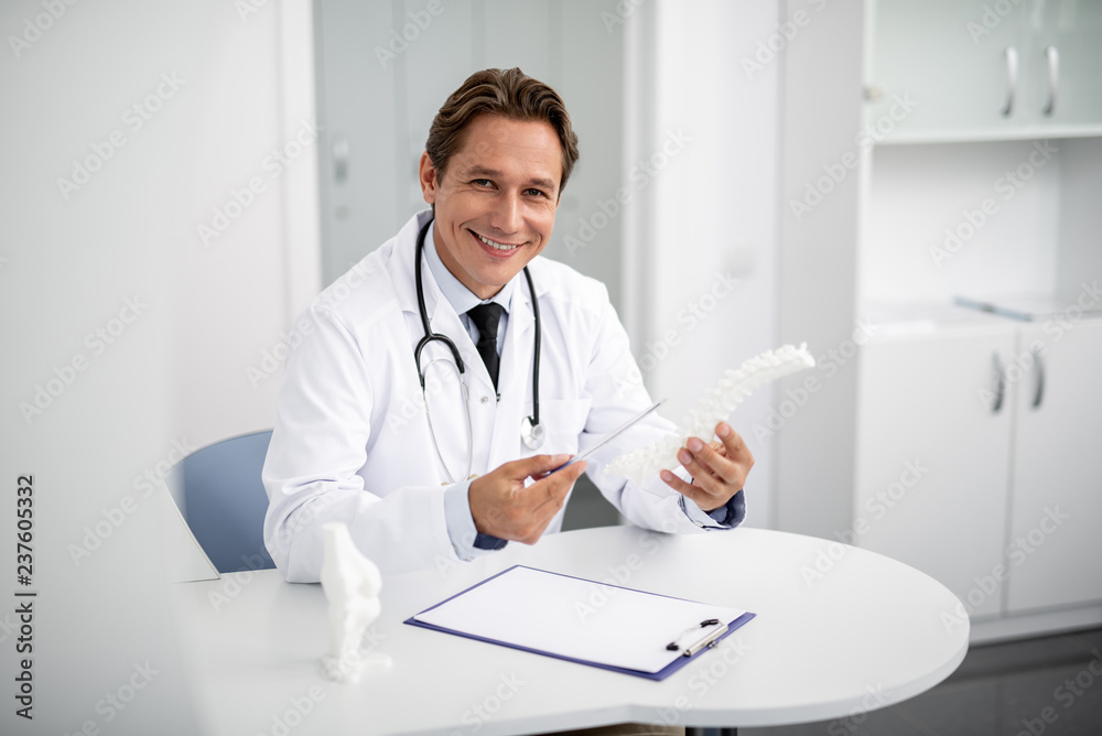 Cheerful general practitioner sitting at the table with spinal model in his hands and smiling while pointing to it Cheerful general practitioner sitting at the table with spinal model in his hands and smiling while pointing to it