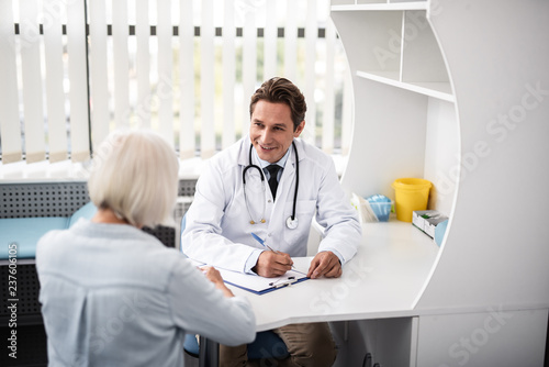 Friendly doctor. Cheerful experienced general practitioner smiling while sitting at the table with his patient and looking at her