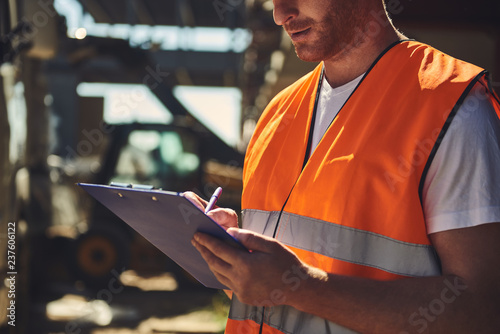 Close up of professional builder in orange uniform standing outdoors and carefully making notes while holding clipboard photo