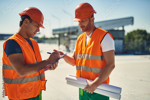 Calm builder in orange helmet and vest standing in front of his colleague and writing on the clipboard
