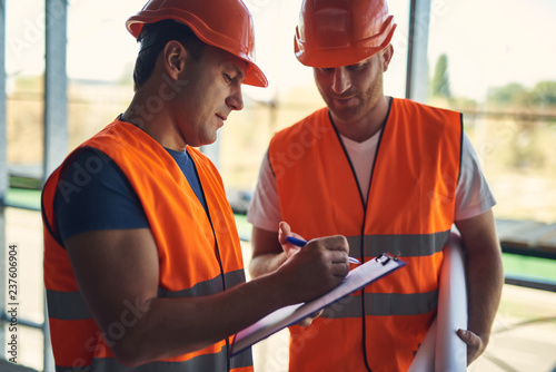 Calm concentrated foreman in the uniform holding clipboard and making notes on the paper while his colleague standing next to him
