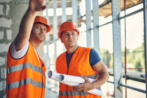 Attentive confident builder pointing to the distance while standing with his reliable coworker in unfinished building