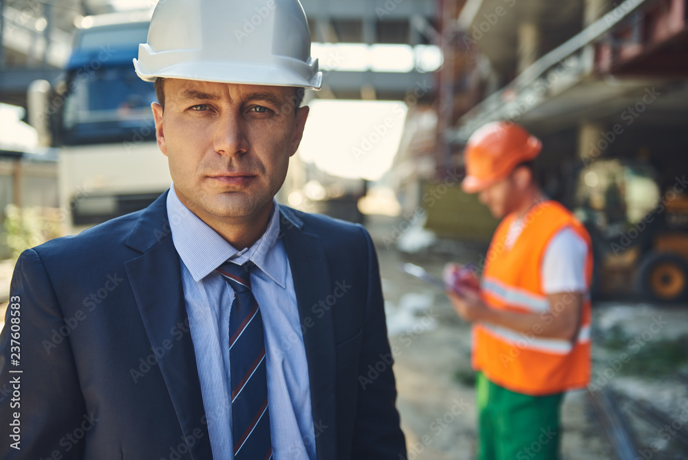 Waist up portrait of adult architect is locating on construction site while male worker standing on background Waist up portrait of adult architect is locating on construction site while male worker standing on background