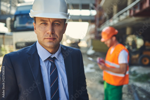 Waist up portrait of adult architect is locating on construction site while male worker standing on background