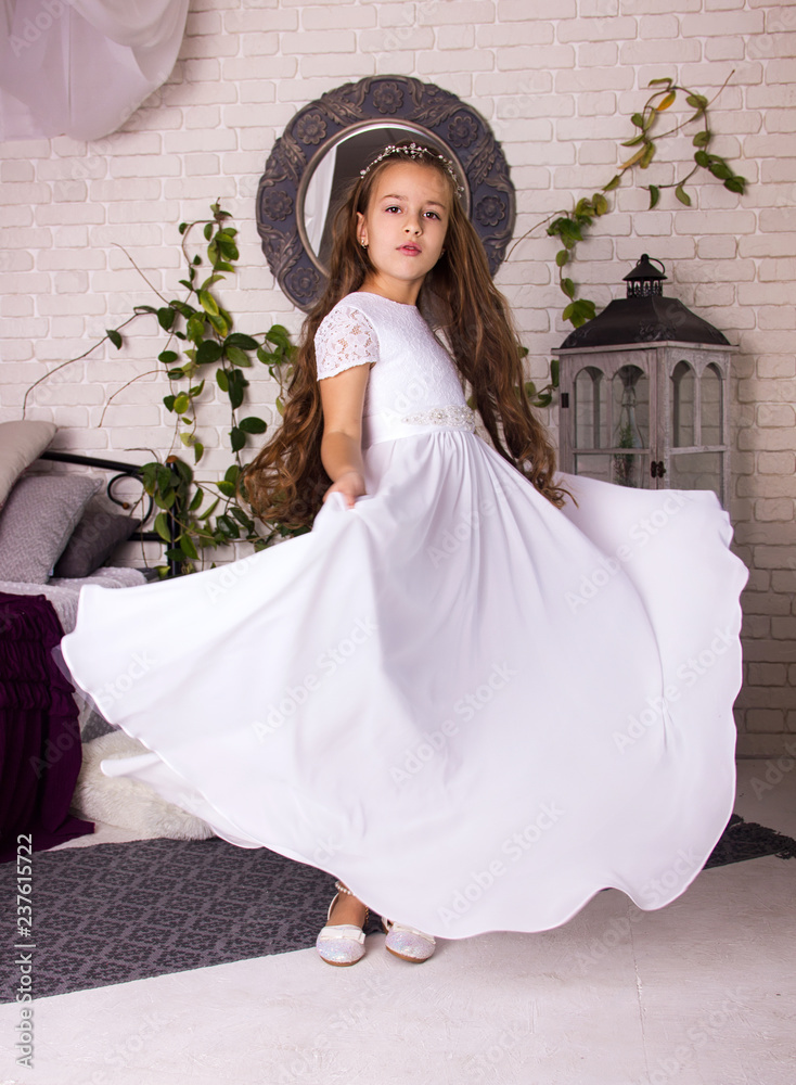 Fototapeta premium little girl in white shiny dress with bow. kid christmas costume. young girl princess in white room is standing from the back like princess in white fancy skirt 