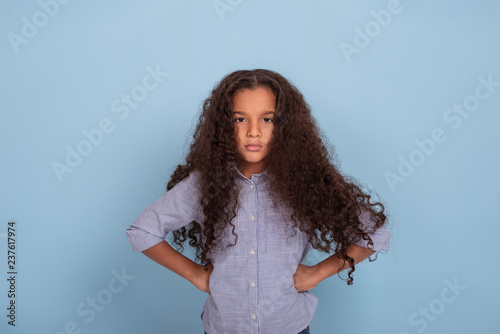 Close up  emotional portrait of frizzy girl wearing blue shirt. She is standing hands in hips and looking angrily at the camera on a blue background in the studio. photo