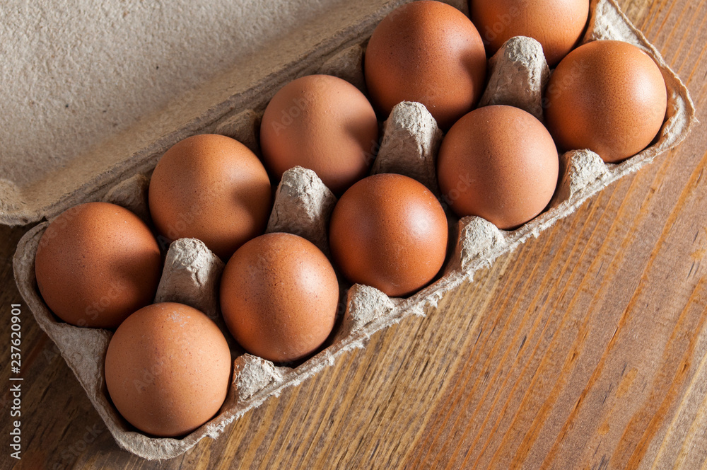 Ten brown chicken eggs lay in a carton on a wooden table. View from above. Ten brown chicken eggs lay in a carton on a wooden table. View from above.