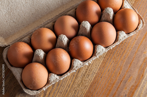 Ten brown chicken eggs lay in a carton on a wooden table. View from above.