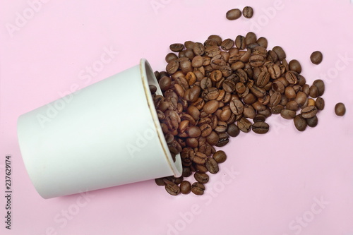 Roasted dark coffee beans are beautifully poured from white paper cups on a table with a pink background isolated