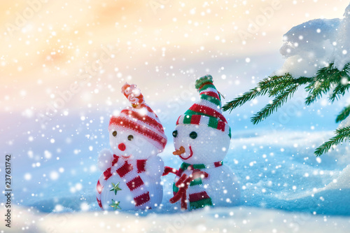 Two small funny toys baby snowman in knitted hats and scarves in deep snow outdoors near pine tree branch. Happy New Year and Merry Christmas greeting card.