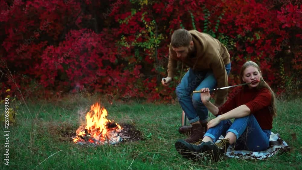 Young man getting up coming towards camera while blonde girl eats fried sausage on colorful autumn forest picnic slowmo Young man getting up coming towards camera while blonde girl eats fried sausage on colorful autumn forest picnic slowmo