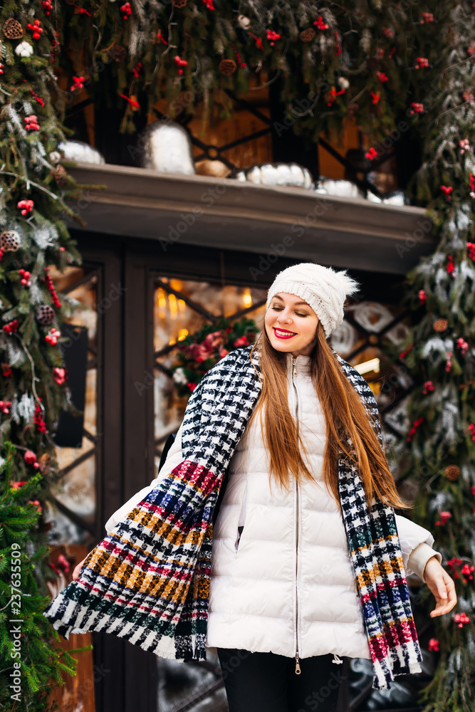 cute girl in winter clothes looks down on the background of a beautiful cafe with glass doors and Christmas decorations.