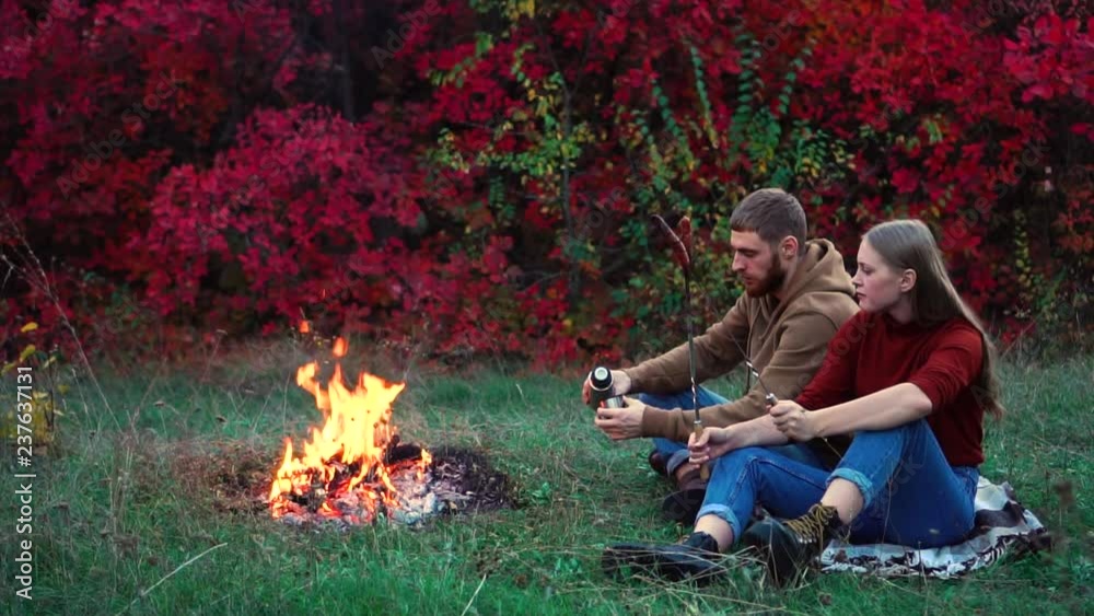 Young friends having calm sausage lunch outdoor on campfire picnic barbecue in colorful autumn red forest in slow motion
