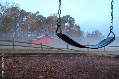 A lonesome melancholic scene of an empty swing with an old red barn and a broken wooden fence on a cool foggy morning. Lake Benson Park in Garner North Carolina. photo