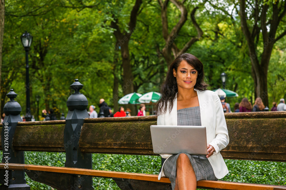 Naklejka premium Young American Woman traveling, working in New York, wearing gray patterned open neck dress, white coat, sitting on long bench at park under green trees, working on laptop computer, looking, thinking