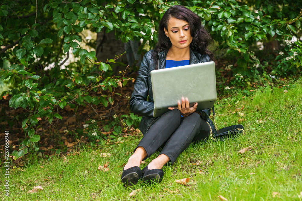 Young American Woman traveling, working in New York, wearing black leather jacket, black pants, shoes, sitting on green grasses law by bushes at park, small bag on ground, reading laptop computer.. Young American Woman traveling, working in New York, wearing black leather jacket, black pants, shoes, sitting on green grasses law by bushes at park, small bag on ground, reading laptop computer..