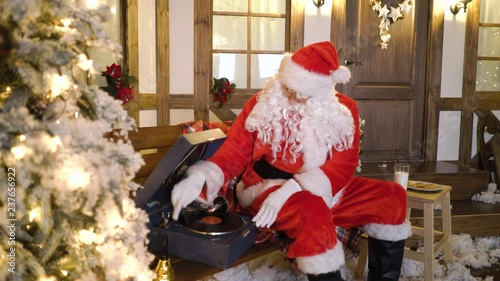 Santa Claus sits near the house between christmas trees, drinks milk, eats cookies, listens christmas songs on vinyl by gramophone, relaxes, enjoys, miss alone