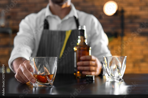 Bartender with glass and bottle of whiskey at counter in bar, closeup. Space for text