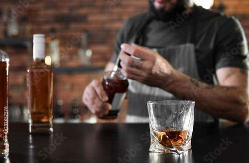 Bartender at counter with bottles and glass of whiskey in bar, closeup. Space for text