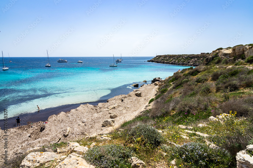 Seascape of Cala Azzurra, a small sandy beach with azure waters and sailboats in the background, Aegadian Islands, Sicily, Italy