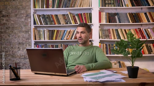 Irritated businessman works on the laptop and makes negative comments. Bookshelves on the background photo