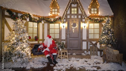 Santa Claus sits near the house between christmas trees, drinks milk, eats cookies, listens christmas songs on vinyl by gramophone, relaxes, enjoys, miss alone