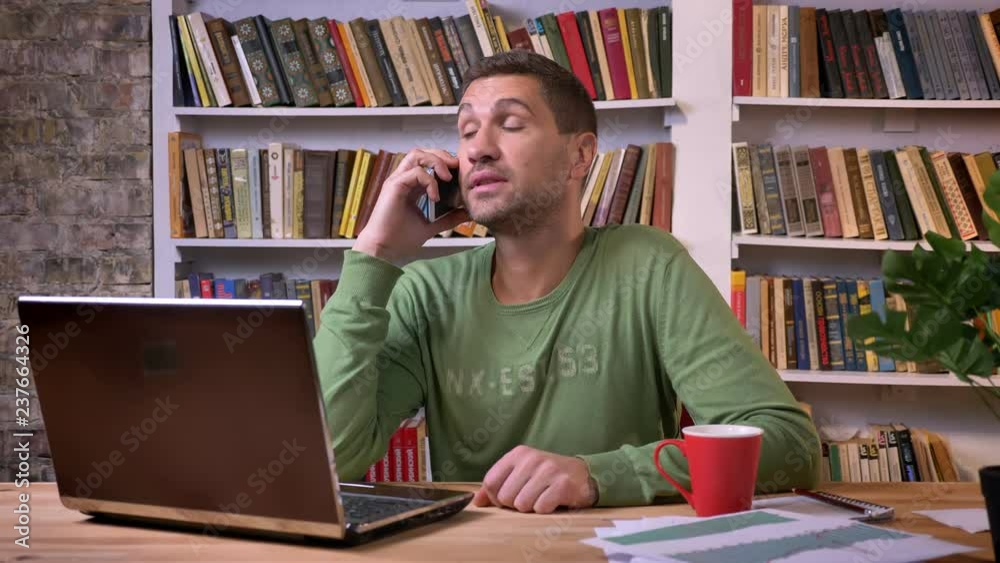 Office worker sitting in front of the laptop and talking on a phone with warm facial expression. Bookshelves on the back