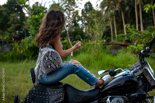 A woman sitting on a bike near fantastic landscape, jungle, tropical forest in front of her and the nature.Concept of style, fashion in adventure and travel with python accessory