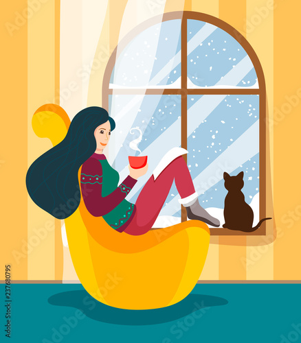 The girl sits at the winter window in a comfortable pillow-chair with a hot cup of drink and reads the story, and a cat sits on the windowsill