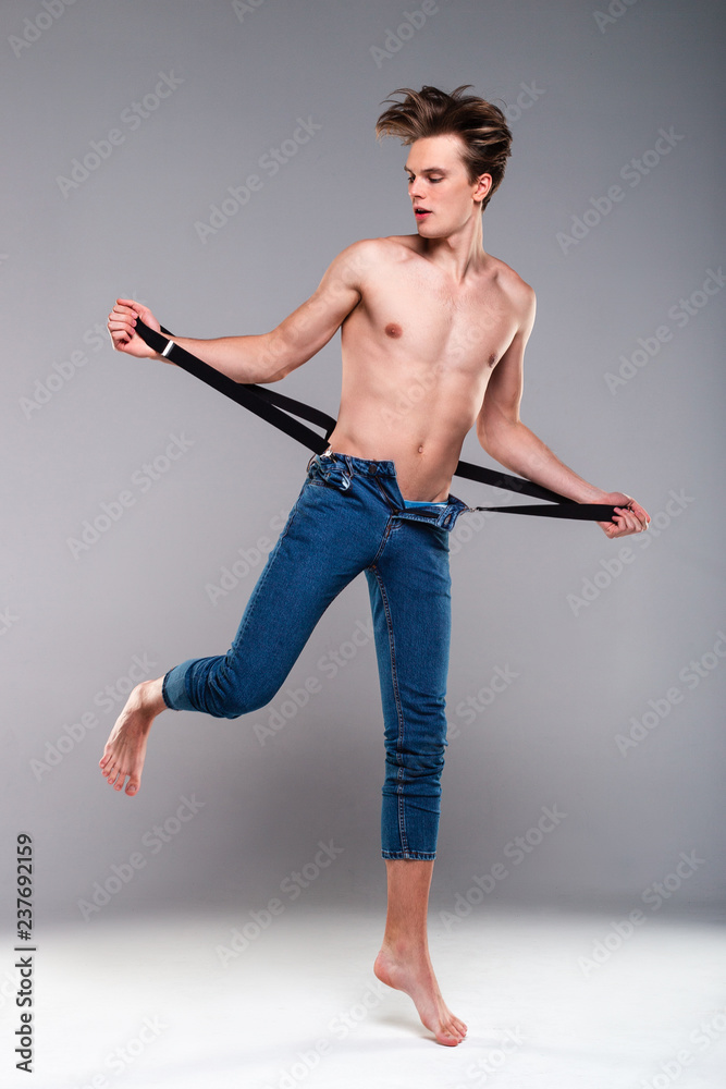 His style. Handsome shirtless young man in jeans holding suspenders and looking away while standing against grey background His style. Handsome shirtless young man in jeans holding suspenders and looking away while standing against grey background