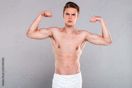 Powerful dude. Handsome shirtless young man wearing towel on his hips showing his biceps and looking at camera while standing against grey background