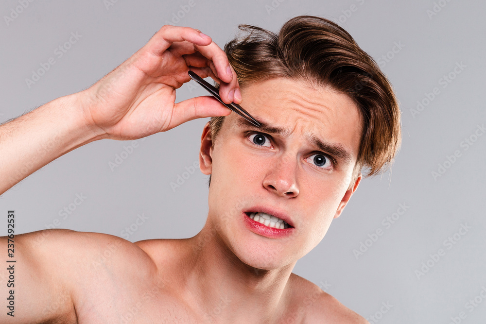 Grooming can be painful. Portrait of handsome shirtless young man tweezing his eyebrows and looking at camera while standing against grey background