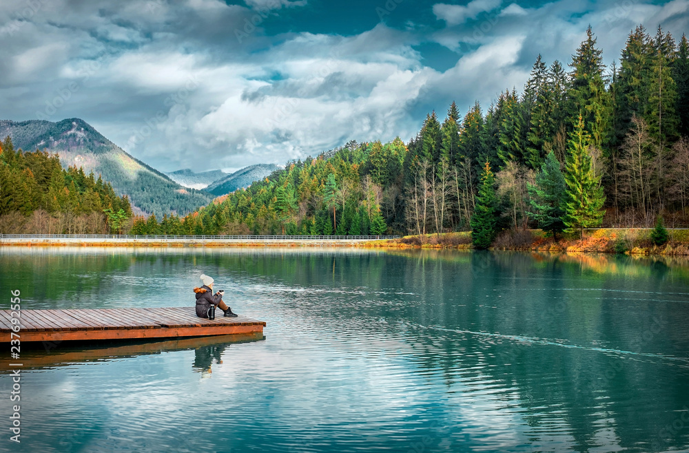 Happy young woman sitting on the wooden pier and drink coffee, lake mountains background Happy young woman sitting on the wooden pier and drink coffee, lake mountains background