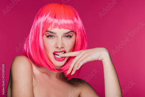 fashionable naked girl in neon pink wig biting finger, isolated on pink
