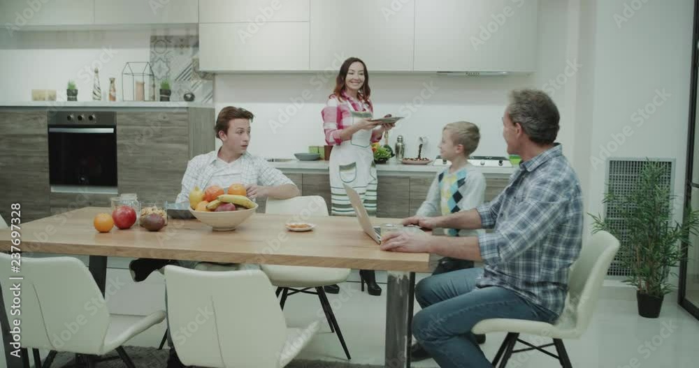 Big family in the morning have a healthy colorful breakfast beautiful mature mom preparing food for her boys and putting on the dinner table dad happy sitting beside and working on his laptop , mature Big family in the morning have a healthy colorful breakfast beautiful mature mom preparing food for her boys and putting on the dinner table dad happy sitting beside and working on his laptop , mature