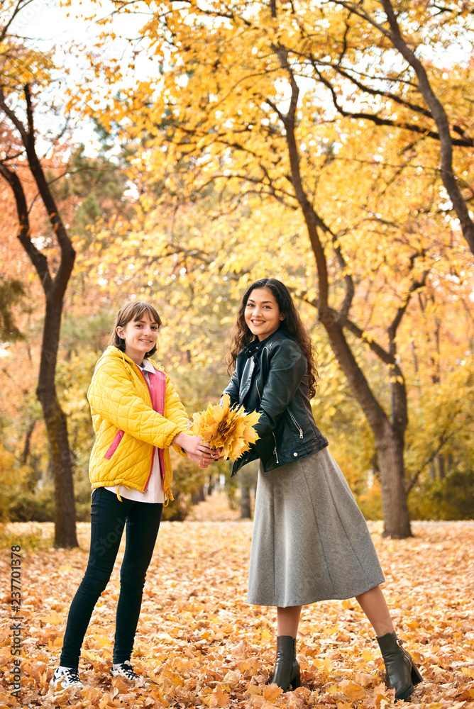 Pretty woman and teen girl are posing with bunch of maple's leaves in autumn park. Beautiful landscape at fall season. Pretty woman and teen girl are posing with bunch of maple's leaves in autumn park. Beautiful landscape at fall season.