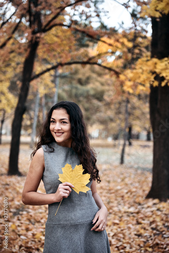 Pretty woman posing with maple's leaf in autumn park near big tree. Beautiful landscape at fall season.