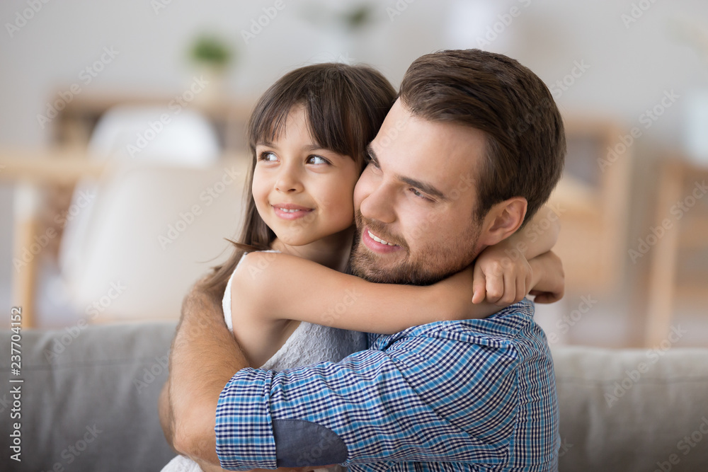Head shot diverse preschool daughter sitting on sofa together with caucasian young father and hugging. Support protection and warm relations between dad and kid, daddy is best friend concept Head shot diverse preschool daughter sitting on sofa together with caucasian young father and hugging. Support protection and warm relations between dad and kid, daddy is best friend concept