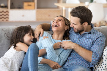 Cheerful people sitting on couch in living room have fun little daughter tickling mother laughing together with parents enjoy free time playing at home. Weekend activity happy family lifestyle concept