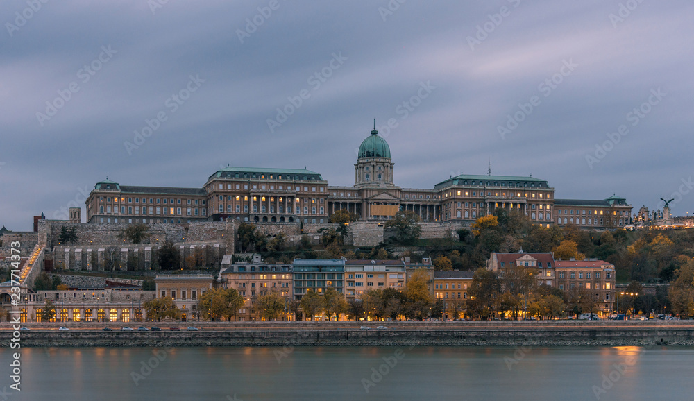 Buda Castle, overlooking the river Danube, in Budapest. It is early evening, and the castle is lit up, with the lights reflecting on the river