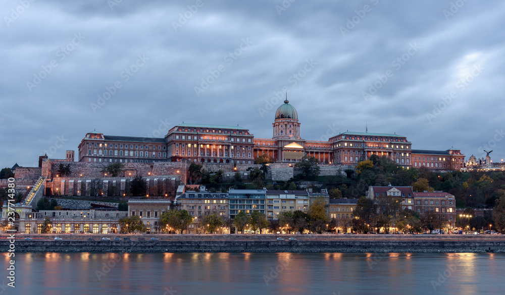 Buda Castle, overlooking the river Danube, in Budapest. It is early evening, and the castle is lit up, with the lights reflecting on the river