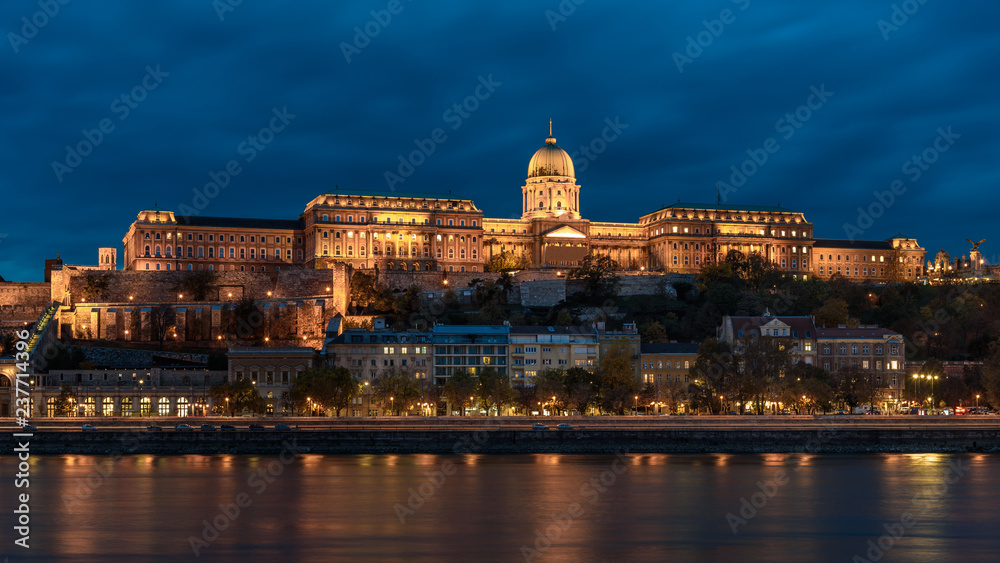 Buda Castle, overlooking the river Danube, in Budapest. The castle is lit up at night time, with the lights reflecting on the river