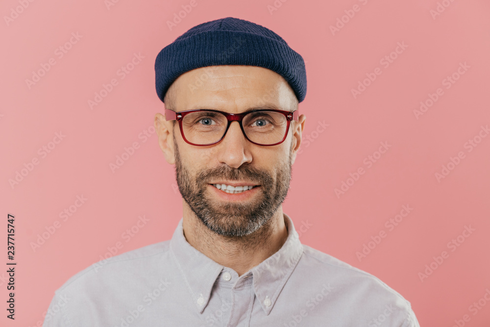 Close up portrait of smiling unshaven male, rejoices good news, wears hat and shirt, looks with eyes full of joy isolated over pink background. European man with bristle ready for date with girlfriend - obrazy, fototapety, plakaty 