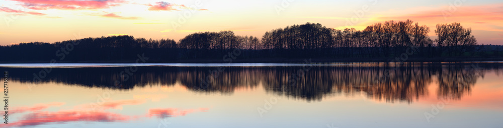 Panorama of a pink and orange sunset soft clouded sky, reflected in the calm water of a rural lake with a silhouetted trees coastline.