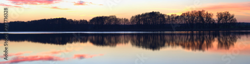 Panorama of a pink and orange sunset soft clouded sky, reflected in the calm water of a rural lake with a silhouetted trees coastline.